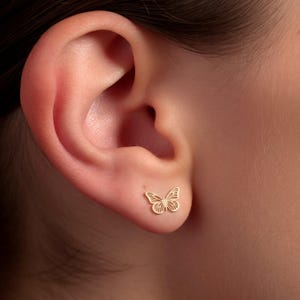 May include: A gold-toned stud earring shaped like a butterfly with intricate, open-work wing details. The small, delicate piece features a symmetrical design with thin lines forming the wing patterns, securely positioned on the lower earlobe.