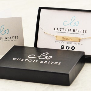 May include: A gold-colored necklace with the name "Rebecca" engraved on a rectangular pendant, presented in a black box with the "Custom Brites" logo. A matching card with the logo is also visible.