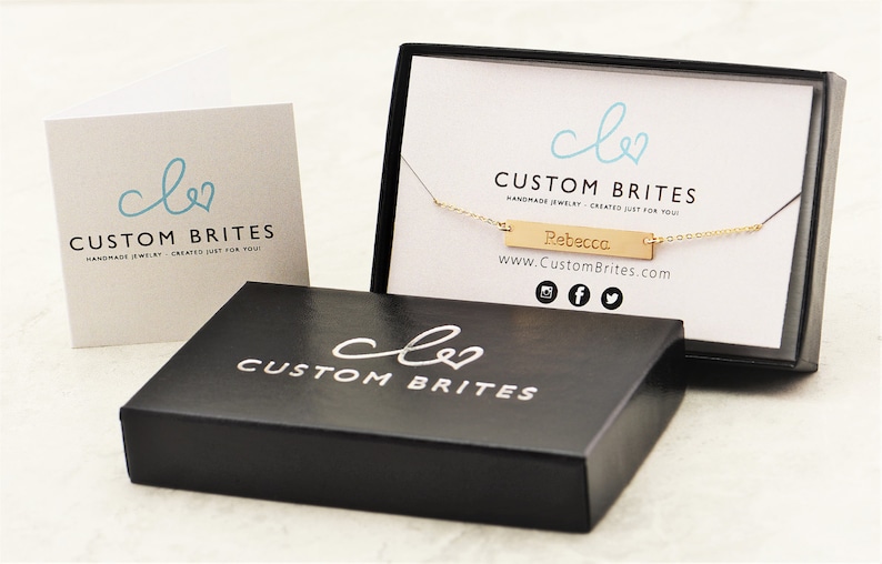 May include: A gold-colored necklace with the name "Rebecca" engraved on a rectangular pendant, presented in a black box with the "Custom Brites" logo. A matching card with the logo is also visible.