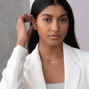 May include: A woman with long black hair wearing a white blazer and a gold necklace with a bar pendant. She is wearing a small gold earring and a nose ring.