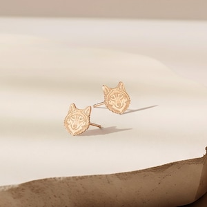 May include: A pair of rose gold stud earrings featuring a detailed engraved wolf head design. Each earring has a post back and displays the facial features of a wolf, including pointed ears and textured fur lines, rendered in a metallic finish.