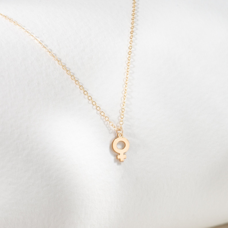 May include: A gold chain necklace with a small, delicate pendant in the shape of the female symbol.