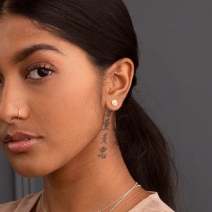 Puede incluir: Una mujer con el pelo oscuro y un piercing en la nariz lleva un peque&ntilde;o pendiente de calavera de oro. Tiene un tatuaje en el cuello con caracteres japoneses.
