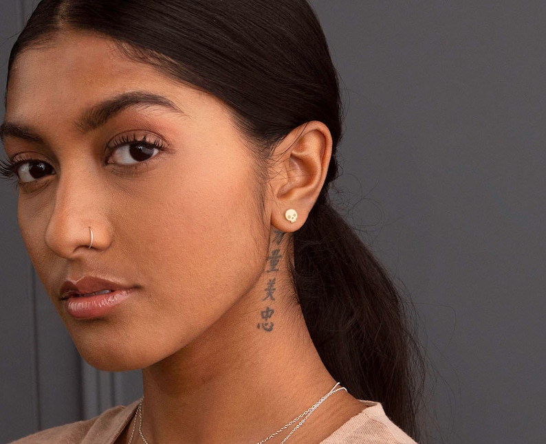 Puede incluir: Una mujer con el pelo oscuro y un piercing en la nariz lleva un peque&ntilde;o pendiente de calavera de oro. Tiene un tatuaje en el cuello con caracteres japoneses.