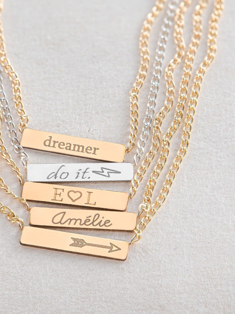 May include: A gold chain necklace with five bar pendants. The pendants are engraved with the words "dreamer", "do it. ", "E&hearts;L", "Am&eacute;lie", and an arrow.