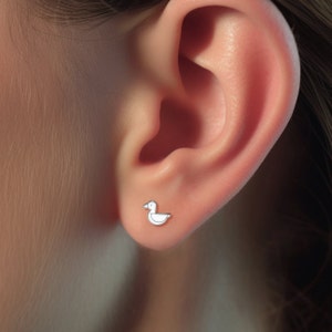 Puede incluir: Primer plano de la oreja de una persona con un peque&ntilde;o pendiente de pato blanco. El pendiente es un dise&ntilde;o sencillo con un cuerpo redondeado y un peque&ntilde;o pico.