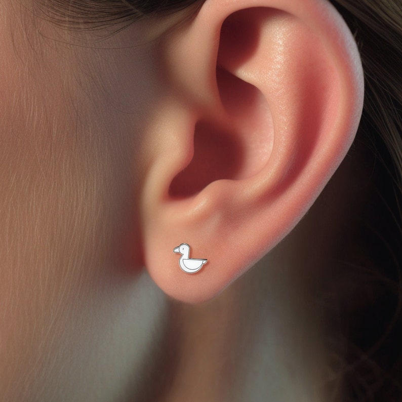 Puede incluir: Primer plano de la oreja de una persona con un peque&ntilde;o pendiente de pato blanco. El pendiente es un dise&ntilde;o sencillo con un cuerpo redondeado y un peque&ntilde;o pico.