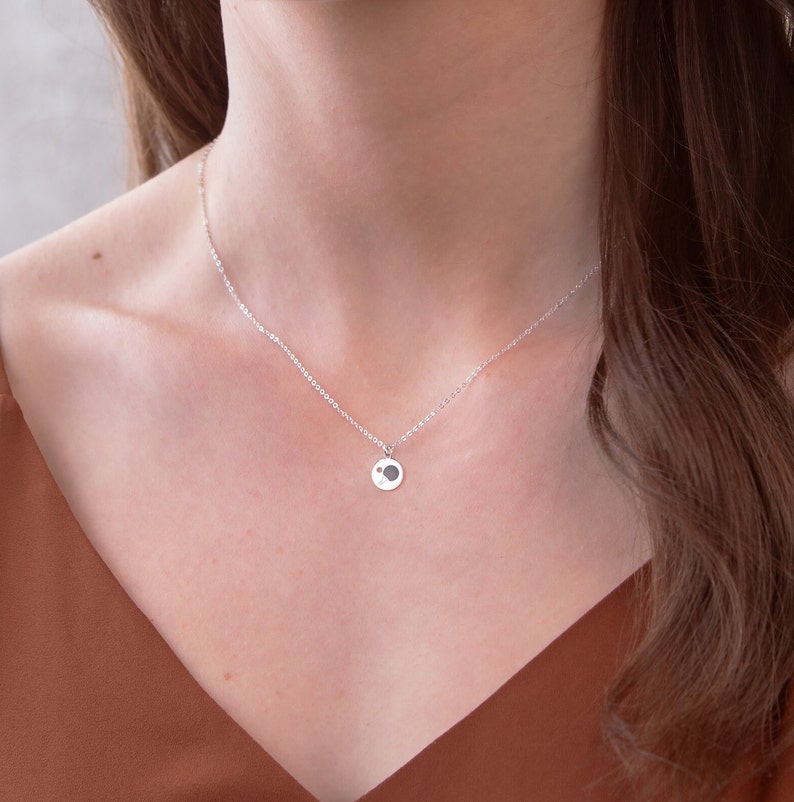 May include: A silver chain necklace with a small, round pendant featuring a minimalist design. The pendant is a circle with a smaller circle cut out of the centre.