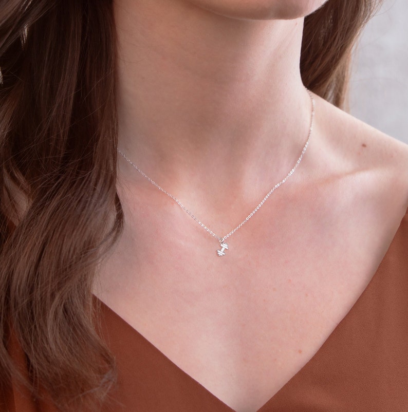 May include: A silver necklace with a small dumbbell pendant. The necklace is worn by a person with brown hair and a brown top.