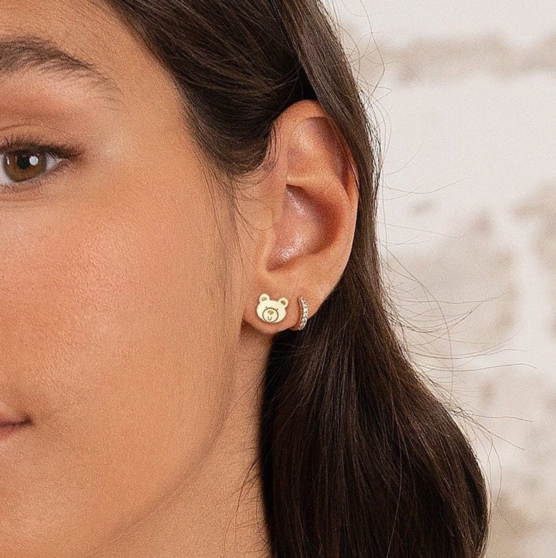 May include: A pair of gold earrings, one featuring a small bear design and the other a small hoop with sparkling stones.