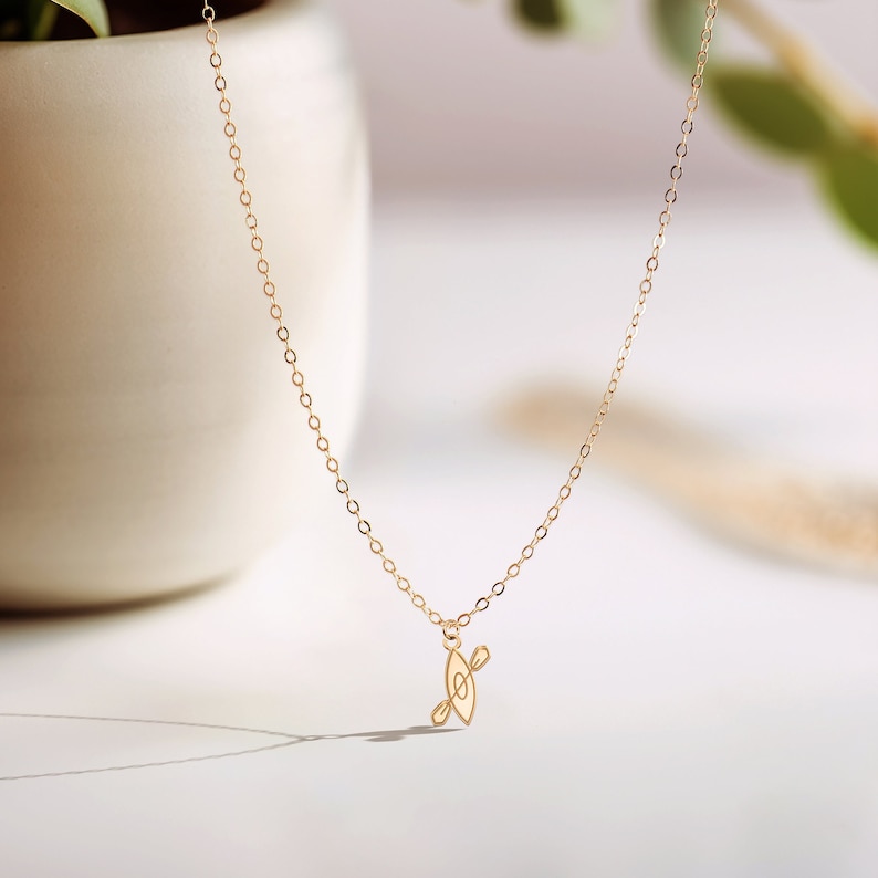 K&ouml;nnte beinhalten: Eine goldene Kettenkette mit einem kleinen, filigranen Anh&auml;nger in Form eines Kanus mit Paddeln. Die Kette liegt auf einem wei&szlig;en Hintergrund.