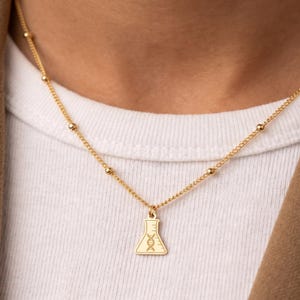 May include: A gold-toned necklace featuring a delicate chain with small evenly spaced beads and a flat pendant shaped like a laboratory Erlenmeyer flask. The flask pendant is engraved with a detailed DNA double helix symbol in the center.