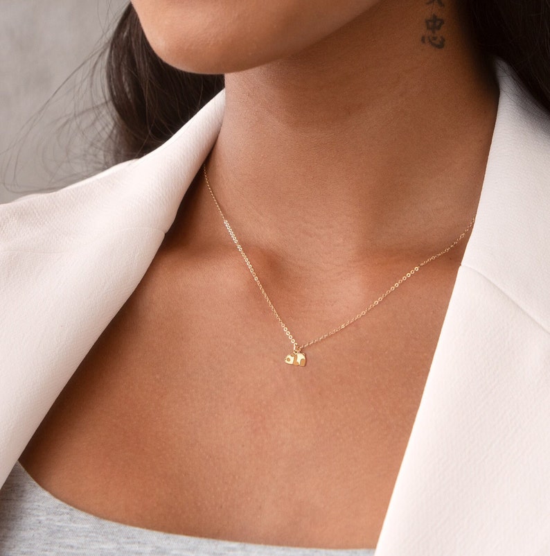 May include: A delicate gold necklace featuring two small, textured pendants. The necklace is suspended from a fine chain and rests against a neutral background, highlighting the jewelry's simple elegance. The pendants appear to be the letters "ah".