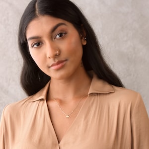 May include: A woman with long dark hair wearing a light brown collared shirt with a gold necklace and a small gold nose ring.