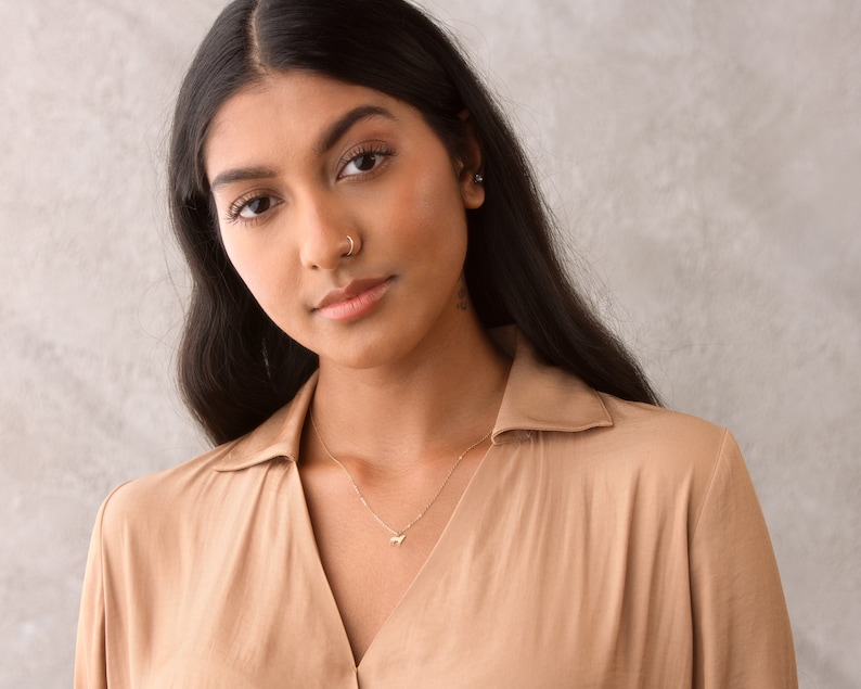 May include: A woman with long dark hair wearing a light brown collared shirt with a gold necklace and a small gold nose ring.