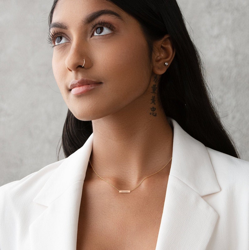May include: A woman wearing a white blazer with a gold bar necklace. She has a nose ring and a small stud earring in her right ear. The necklace has a small bar pendant.
