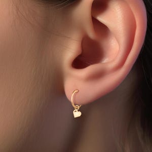 May include: A gold hoop earring with a small heart charm dangling from it. The earring is worn in the upper lobe of a person's ear.
