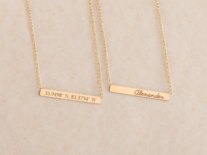 May include: Two gold necklaces with bar pendants. One pendant is engraved with the coordinates "33.9498&deg; N, 83.3734&deg; W". The other pendant is engraved with the name "Alexander".