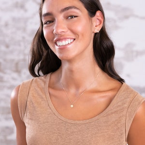 May include: A woman with long brown hair is wearing a light brown sleeveless shirt and a delicate gold necklace with a small round pendant. She is smiling and looking at the camera.