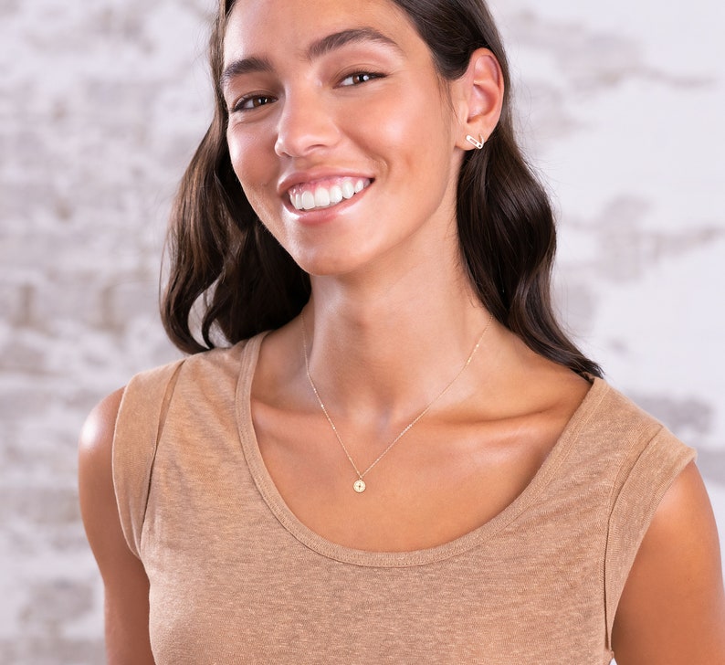 May include: A woman with long brown hair is wearing a light brown sleeveless shirt and a delicate gold necklace with a small round pendant. She is smiling and looking at the camera.
