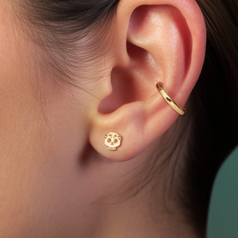 May include: Gold skull stud earrings with a floral design. The earrings are worn in the earlobe.