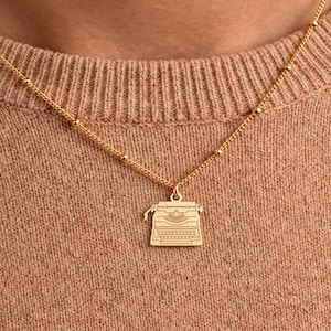 May include: A gold-toned necklace featuring a delicate chain with small evenly spaced beads and a flat, engraved pendant shaped like a vintage typewriter. The pendant displays detailed lines representing the keys and carriage of the machine.