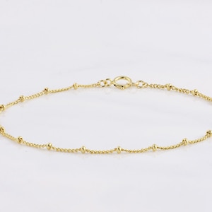 May include: A delicate gold anklet featuring a fine chain interspersed with small, round beads. The anklet has a simple, elegant design with a secure clasp. The jewelry is displayed on a white background.