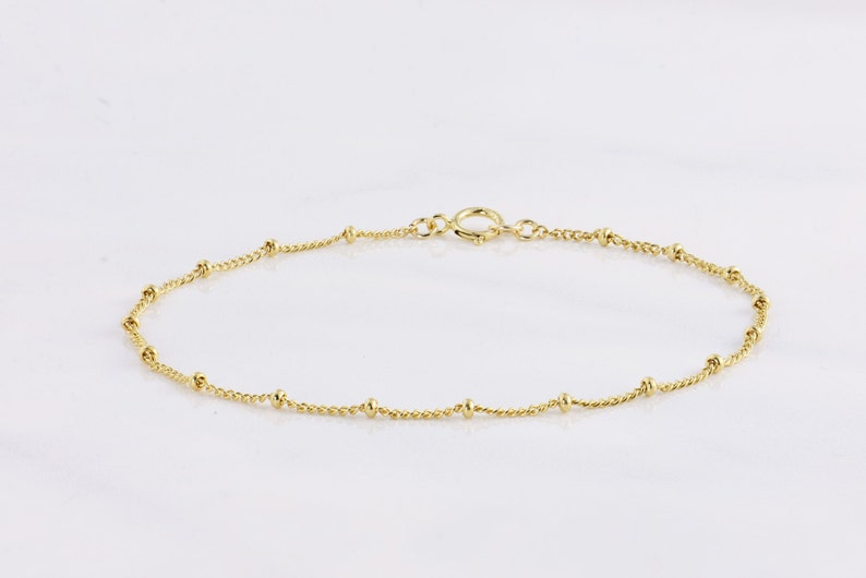 May include: A delicate gold anklet featuring a fine chain interspersed with small, round beads. The anklet has a simple, elegant design with a secure clasp. The jewelry is displayed on a white background.