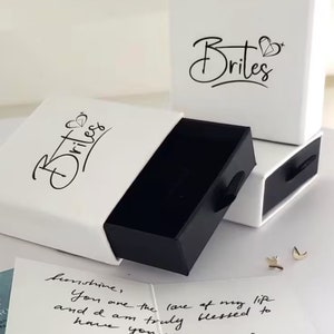 May include: Two white boxes with black lids, one open and one closed. The boxes have the word "Brites" printed on them in black cursive lettering. The open box is revealing a black interior. A handwritten note is visible in the bottom left corner of the image.