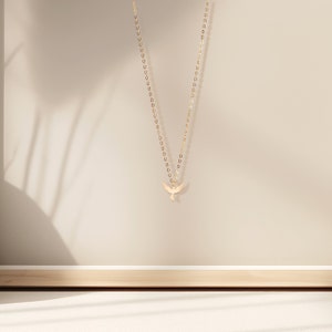 May include: A gold chain necklace with a delicate bird pendant. The necklace is hanging against a beige background.