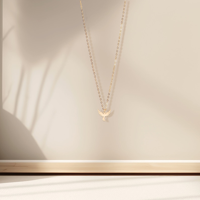 May include: A gold chain necklace with a delicate bird pendant. The necklace is hanging against a beige background.