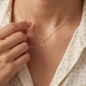Könnte beinhalten: Eine goldfarbene Halskette mit einer feinen Kette und einem kleinen Anhänger in Form von Pfeil und Bogen. Der Anhänger liegt mittig auf dem Schlüsselbein und ist an einer dünnen, polierten Metallgliederkette befestigt, die auf der Haut aufliegt.
