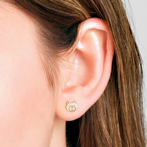May include: A close-up of a person's ear with a small gold bear stud earring. The bear has a simple face with two eyes, a nose, and a mouth.