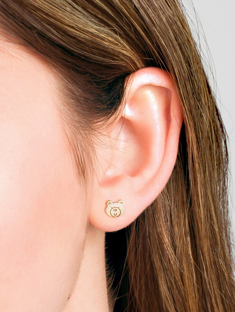 May include: A close-up of a person's ear with a small gold bear stud earring. The bear has a simple face with two eyes, a nose, and a mouth.