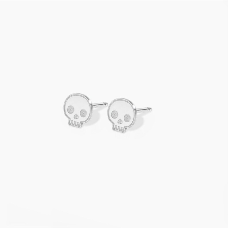 Puede incluir: Un par de pendientes de bot&oacute;n de calavera de plata. Las calaveras son blancas con ojos negros.