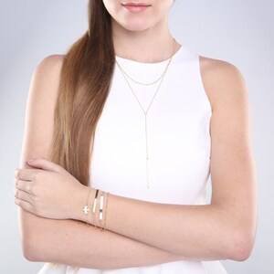 May include: A collection of gold-toned jewelry, including layered necklaces and bracelets. The necklaces feature delicate chains and a Y-shaped pendant. The bracelets include a bar bracelet, a chain bracelet, and a bracelet with a bird charm. The jewelry is displayed on a person wearing a white top.
