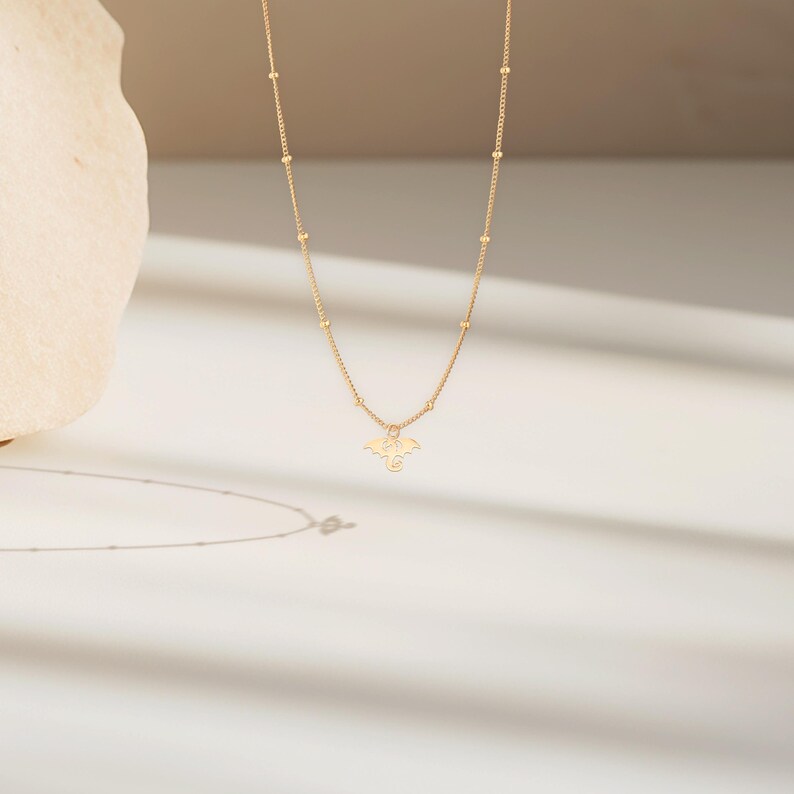 May include: A gold necklace featuring a delicate dragon pendant. The necklace is crafted from tiny gold beads and the dragon is facing forward with its wings outstretched.
