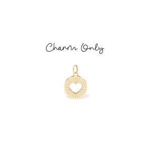 Puede incluir: Un dije dorado con un recorte en forma de corazón en el centro. El dije circular tiene un diseño de rayos de sol. El texto "Charm Only" está escrito encima del dije.