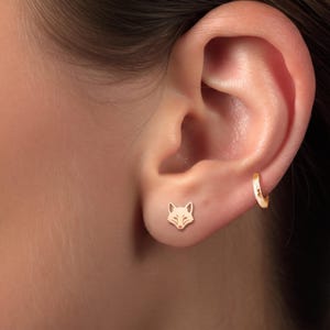 May include: A gold-toned stud earring shaped like a fox head positioned on the earlobe, accompanied by a small, plain gold-toned hoop earring worn in the upper cartilage area of the ear, both featuring a polished metallic finish.
