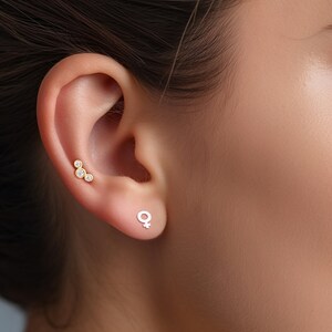 May include: A pair of earrings, one with a cluster of three small round clear stones and the other with a simple gold female symbol.
