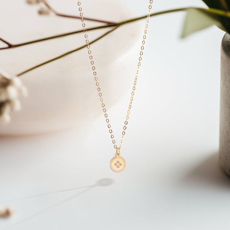 K&ouml;nnte beinhalten: Eine goldene Kettenkette mit einem kleinen, runden Anh&auml;nger, der vier winzige Diamanten in Knopfform tr&auml;gt.