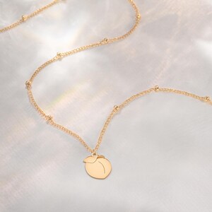 May include: A gold-coloured necklace with a peach-shaped pendant. The chain is dotted with small beads. The necklace is presented on a soft, white background, creating a minimalist aesthetic.