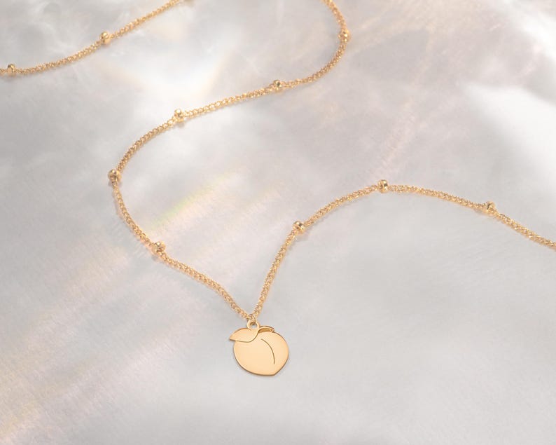 May include: A gold-coloured necklace with a peach-shaped pendant. The chain is dotted with small beads. The necklace is presented on a soft, white background, creating a minimalist aesthetic.