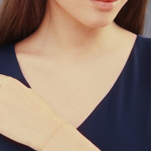 May include: A delicate gold bracelet with small, evenly spaced beads is worn on a wrist. The bracelet is set against a dark blue v-neck top. The skin tone is fair, and the overall aesthetic is minimalist and elegant.