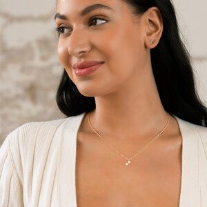 May include: A gold necklace with a small dumbbell pendant. The necklace is worn by a person with dark hair and a white sweater.