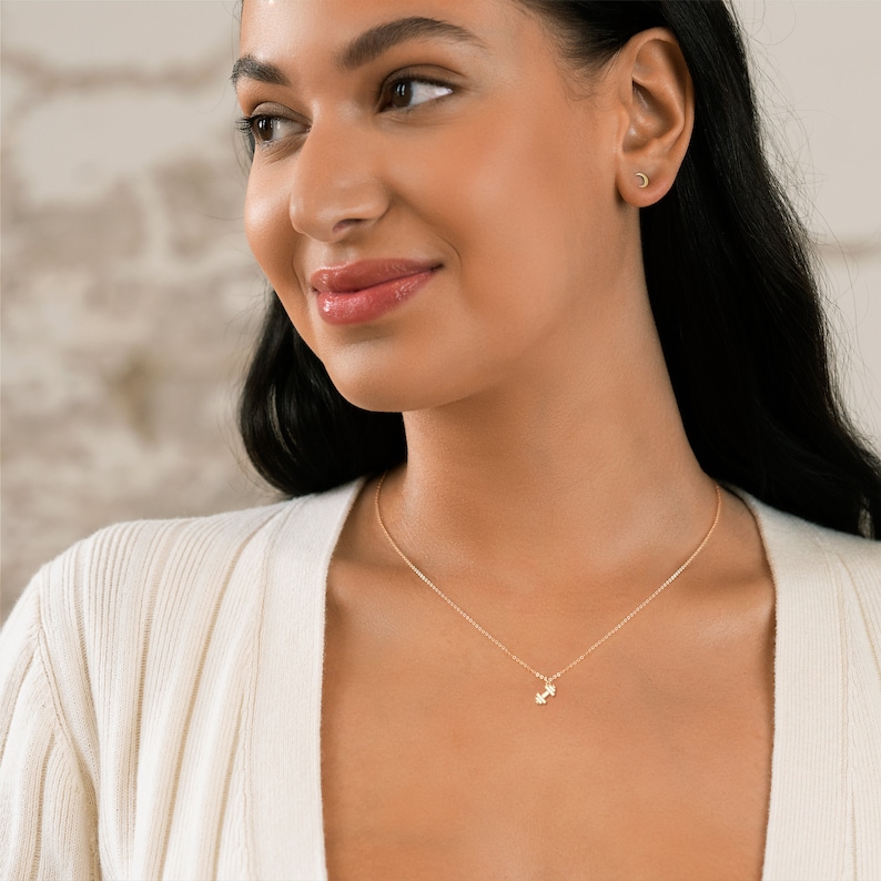 May include: A gold necklace with a small dumbbell pendant. The necklace is worn by a person with dark hair and a white sweater.