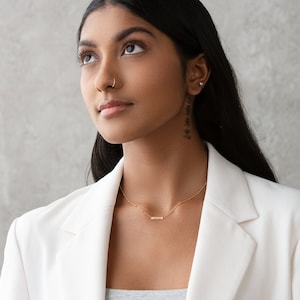 May include: A woman with long dark hair wearing a white blazer and a gold necklace with a small bar pendant. She is looking up and has a nose ring.