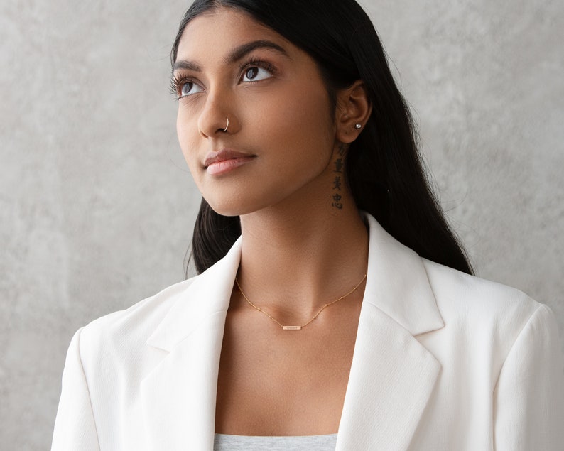 May include: A woman with long dark hair wearing a white blazer and a gold necklace with a small bar pendant. She is looking up and has a nose ring.