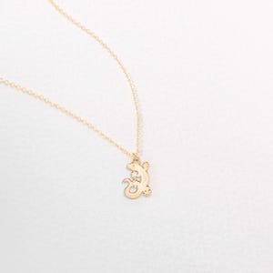 Può includere: Una collana color oro con un ciondolo a forma di lucertola. La lucertola è una silhouette dettagliata con un corpo strutturato, sospesa da una delicata catena. Il gioiello è esposto su uno sfondo bianco.
