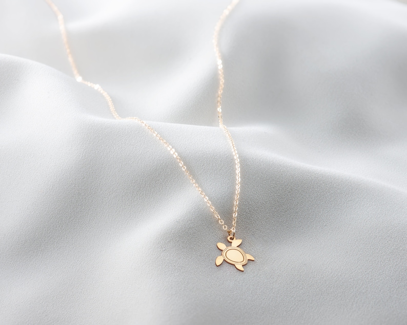 May include: A gold chain necklace with a small turtle pendant. The necklace is on a white fabric background.